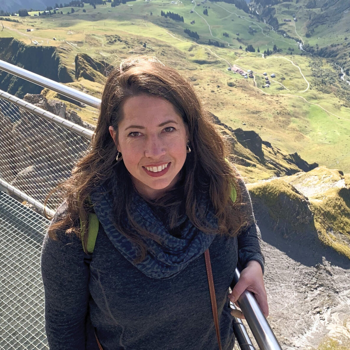 White woman with brown hair smiling at camera posed with hand on guardrail while hiking with mountain vista in background